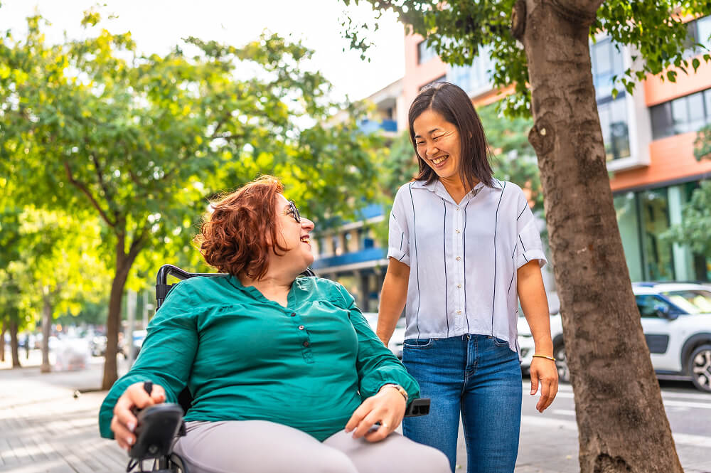An able-bodied woman and a woman in a wheelchair smiling at each other on a street