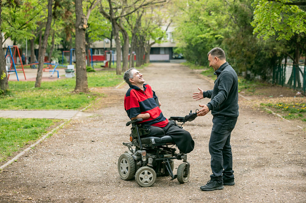 A man in a wheelchair laughing with a man standing in front of him with his arms open, smiling