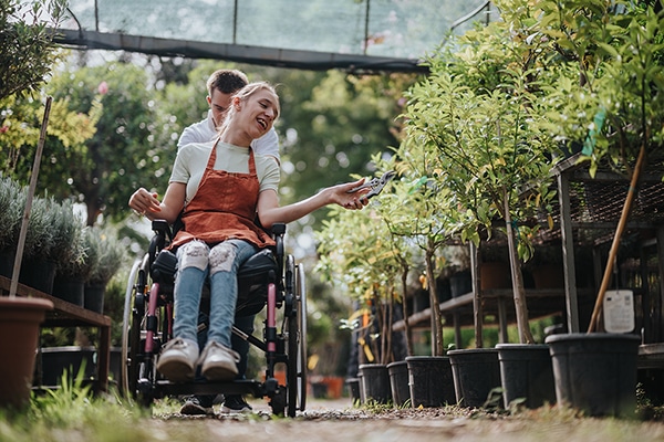 Two friends engage in a joyful gardening activity. A person in a wheelchair enthusiastically tends to plants, symbolising teamwork and inclusivity in a vibrant greenhouse environment.