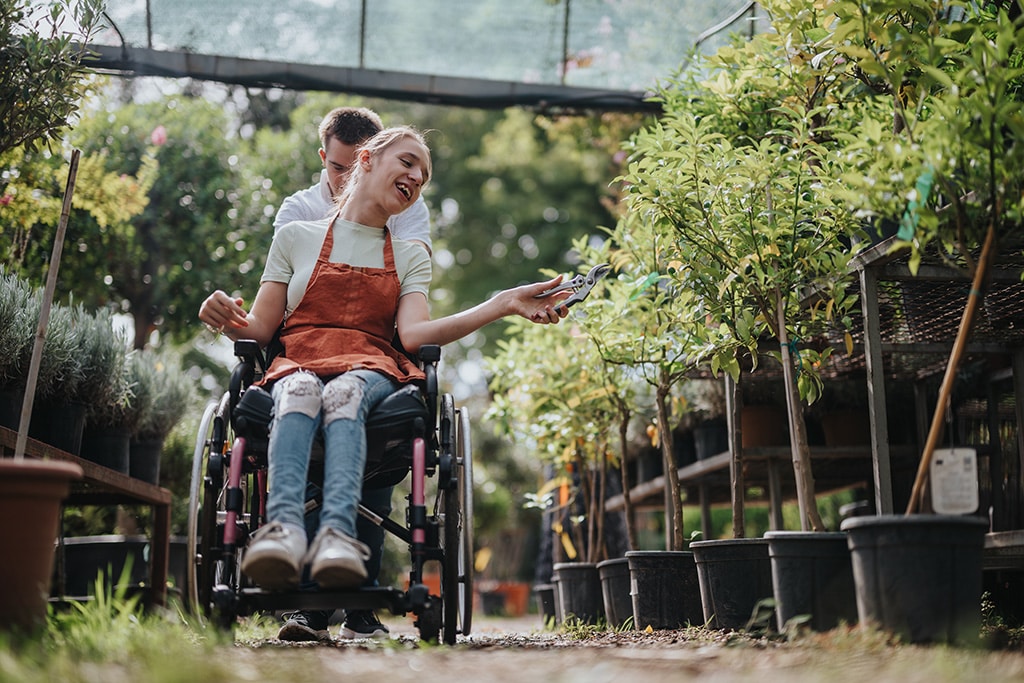 Two friends engage in a joyful gardening activity. A person in a wheelchair enthusiastically tends to plants, symbolising teamwork and inclusivity in a vibrant greenhouse environment.