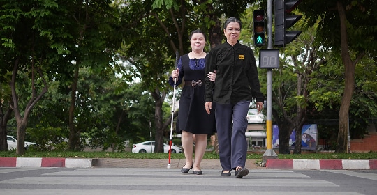 A blind woman crossing the street with the assistance of a carer