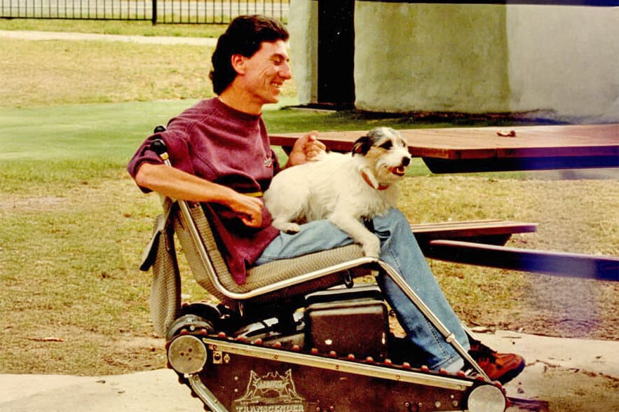 A man in a wheelchair smiling with his dog on his lap, going for a ride