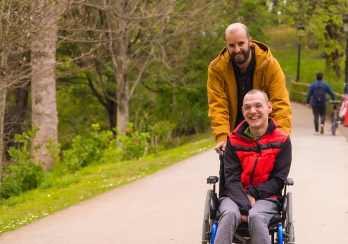A man assisting another man in a wheelchair. They are both smiling.