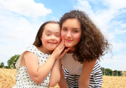 Two young women smiling to camera. One of the young women has Downs Syndrome.