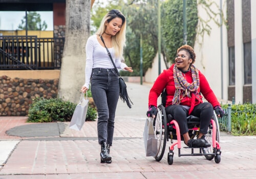 Two women on the pavement. One is wheeling herself in her wheelchair while talking to a woman walking beside her