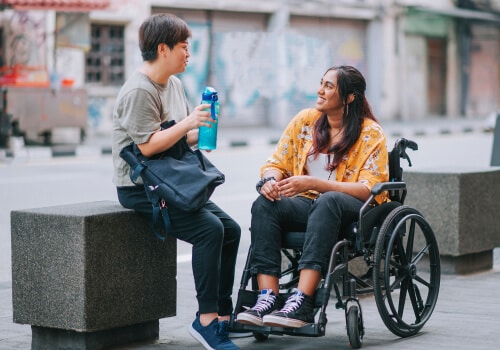 Two people smiling in conversation. One of them sits in a wheelchair while the other perches on a wall