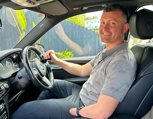 A man sitting in the driver's seat of a car, smiling and holding the steering wheel.