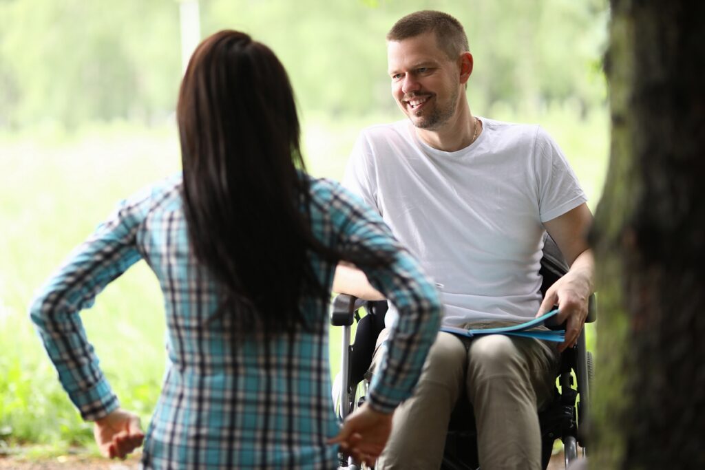 A man in a wheelchair smiles at a woman standing with her back to the camera, outside in a green, natural setting.
