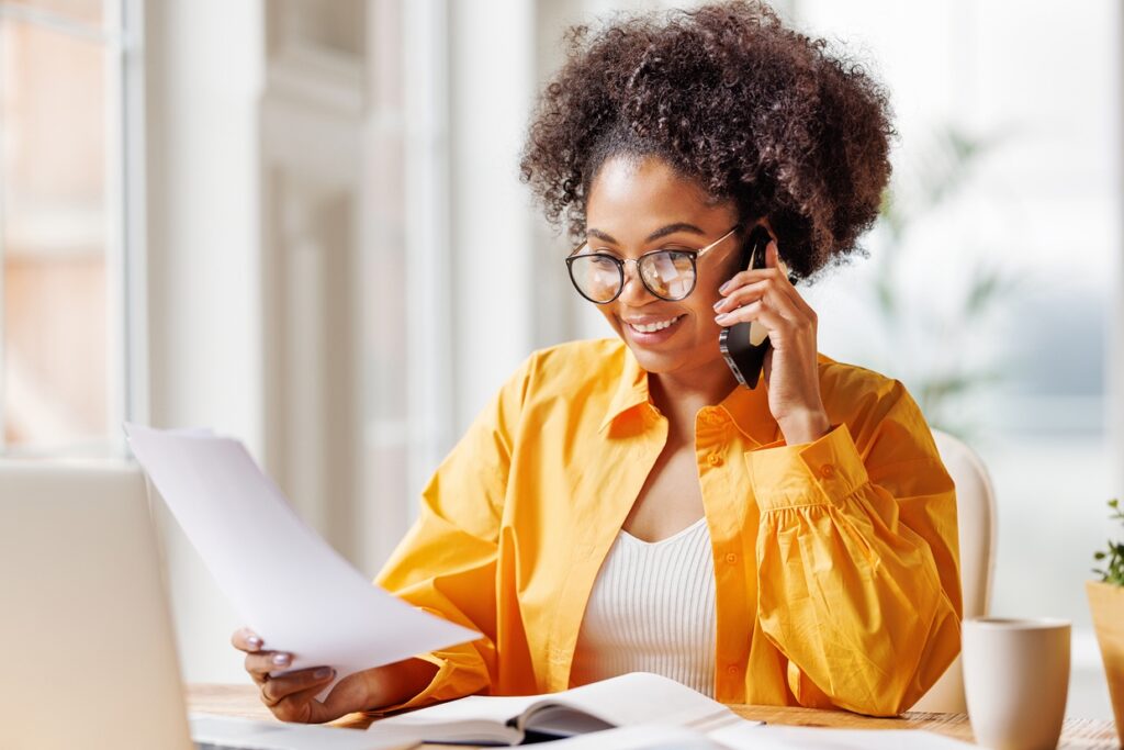 A woman with curly hair is sitting at a desk, smiling while talking on a smartphone. She is holding papers in one hand and has a laptop and a cup of coffee on the table.