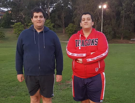 Two young men standing on a grassy field, one wearing a blue hoodie and shorts, the other in a red sports jacket with "Dragons" written on it.