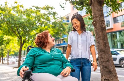 An able-bodied woman and a woman in a wheelchair smiling at each other on a street