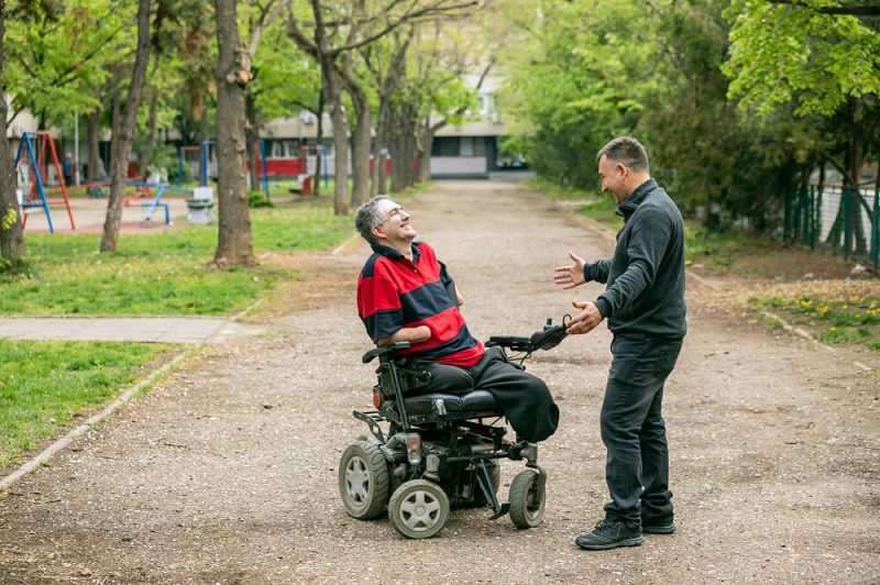 A man in a wheelchair laughing with a man standing in front of him with his arms open, smiling