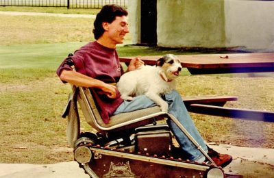 A man in a wheelchair smiling with his dog on his lap, going for a ride