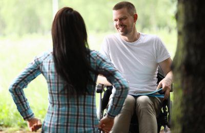 A man in a wheelchair smiles at a woman standing with her back to the camera, outside in a green, natural setting.