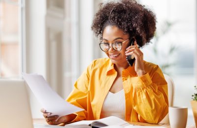 A woman with curly hair is sitting at a desk, smiling while talking on a smartphone. She is holding papers in one hand and has a laptop and a cup of coffee on the table.
