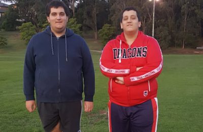 Two young men standing on a grassy field, one wearing a blue hoodie and shorts, the other in a red sports jacket with "Dragons" written on it.